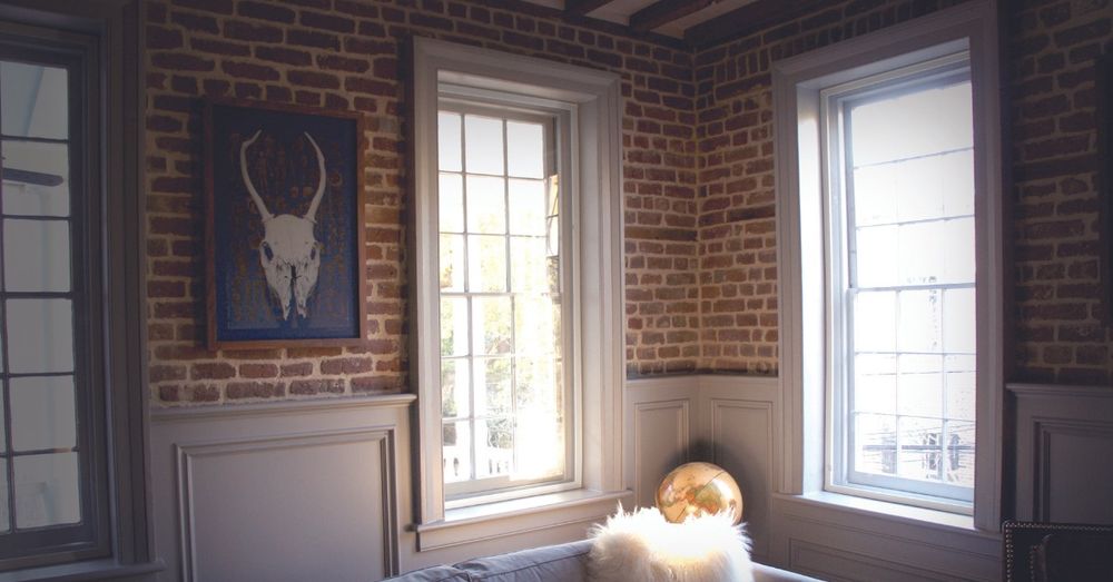 A cozy living room with brick walls, and three large windows that bring
		light into the space. 