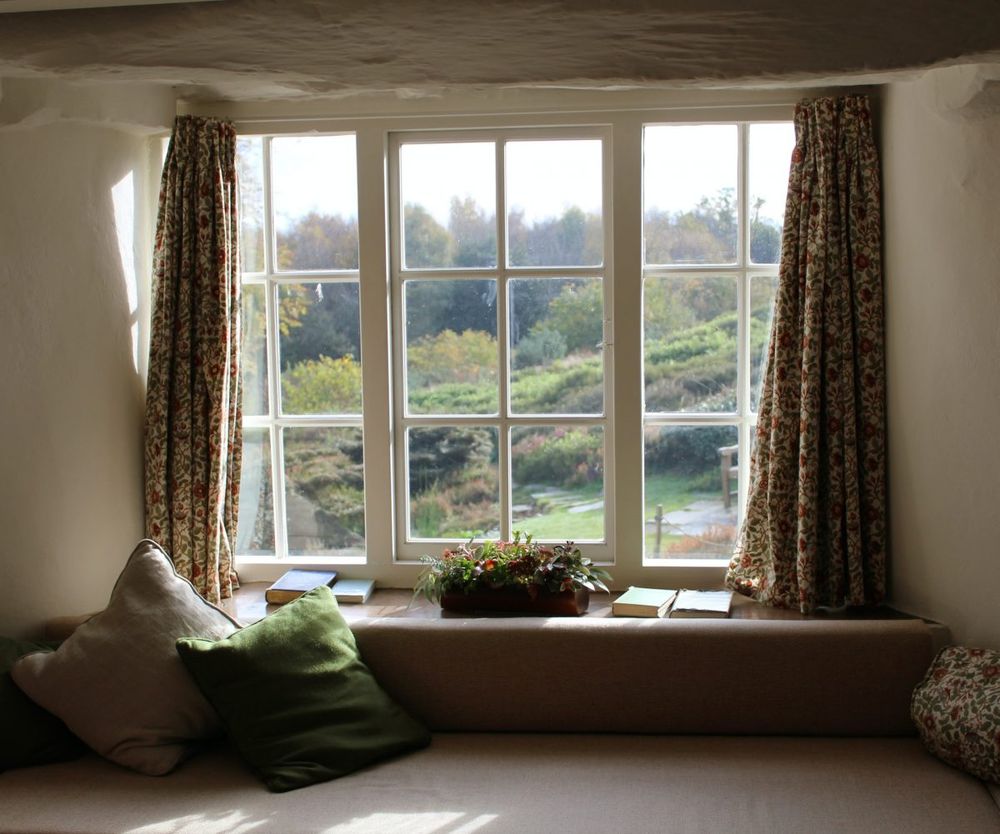 A second-story room with an old window looking out to a backyard. There are curtains hanging from the sides and a ledge by the window holding a plant and some books.